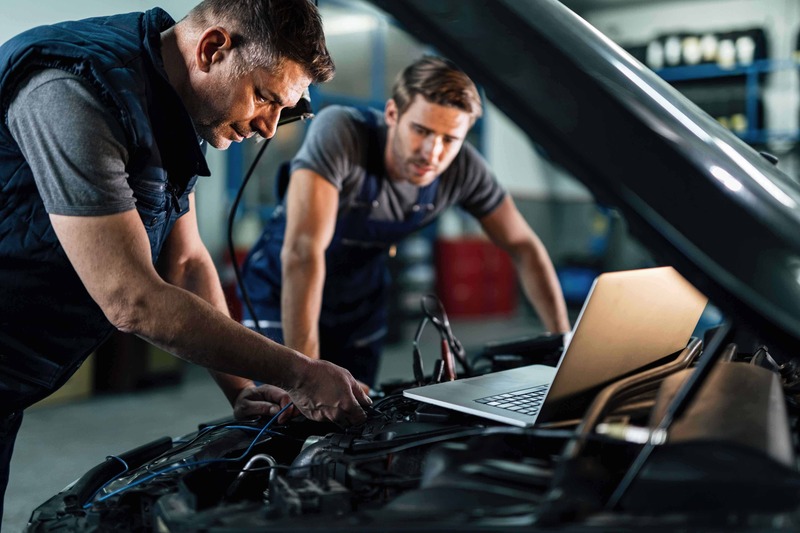 Un mecanic in prim plan verifica masina sub capota, in timpul la verificare itp. Un alt mecanic in fundal priveste si invata